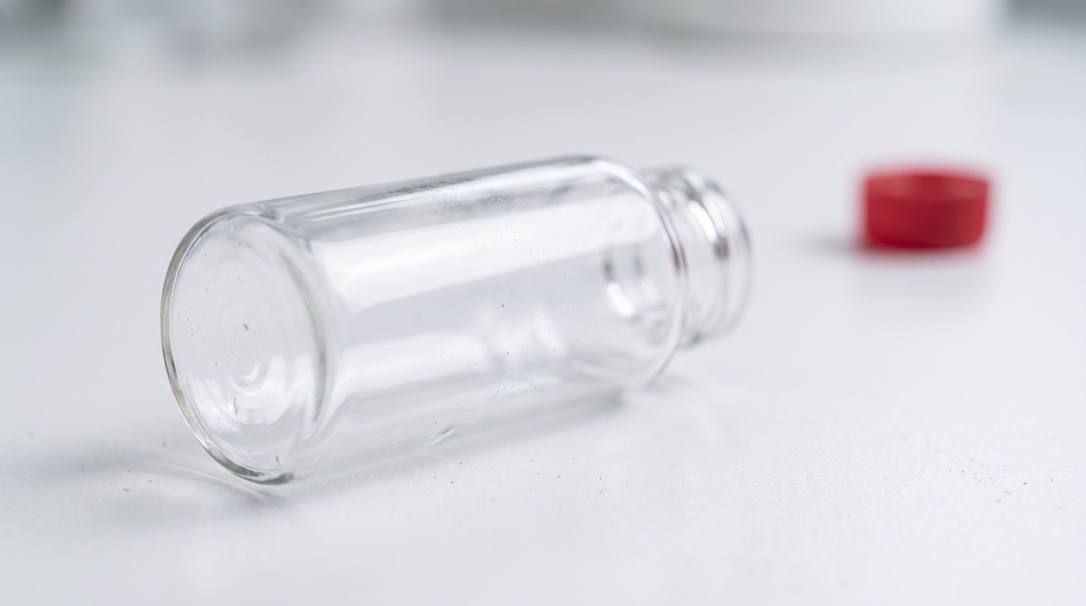 Vial de laboratorio sin etiqueta sobre superficie blanca con acento rojo al fondo